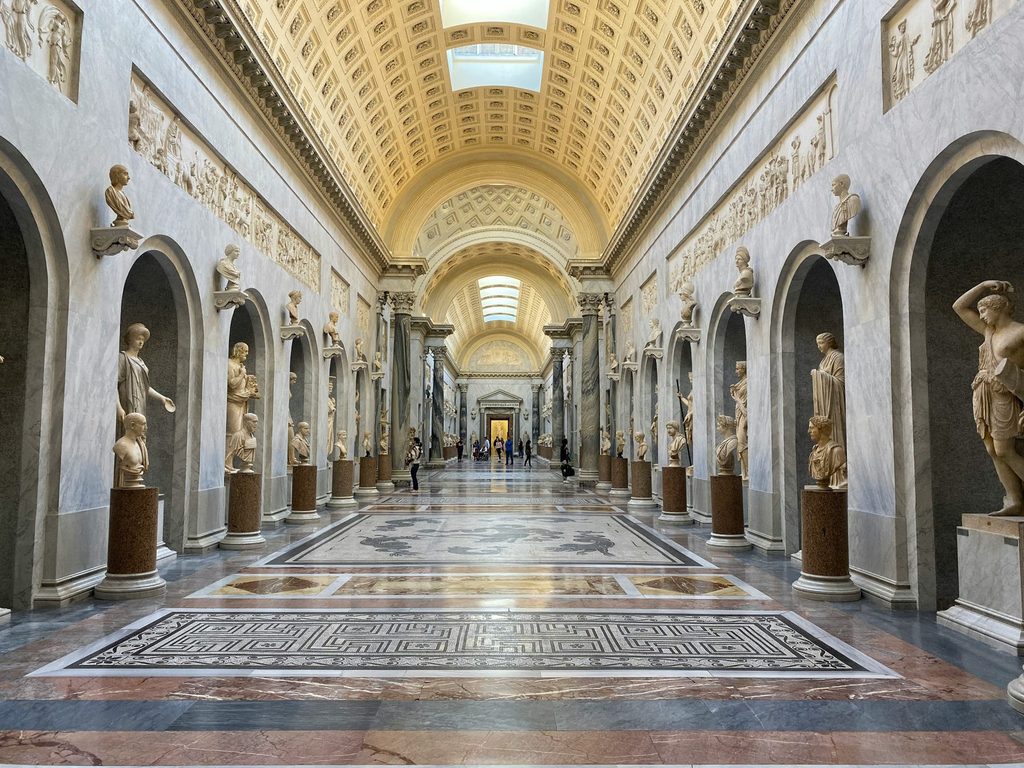 Interior gallery of the Vatican Museums in Rome, featuring classical sculptures, marble floors and an ornate vaulted ceiling.