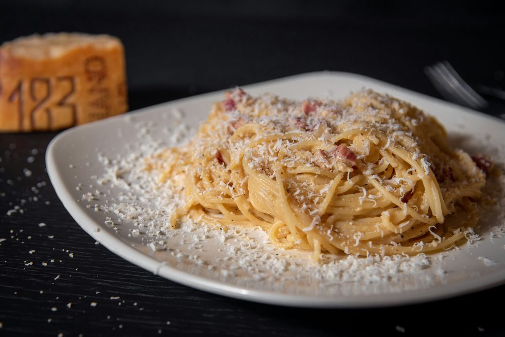 Plate of spaghetti carbonara with grated cheese, black pepper and pieces of cured pork on a dark restaurant table.