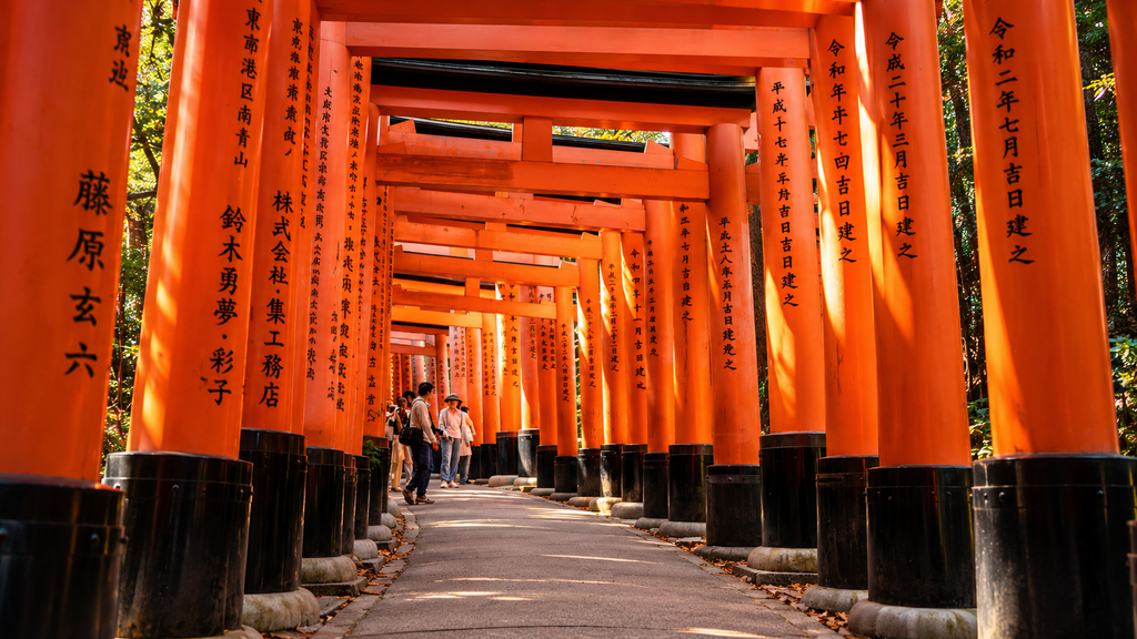 Vermilion torii gates forming a winding pathway at Fushimi Inari Taisha Shrine in Kyoto, Japan, with Japanese inscriptions and visitors walking beneath the gates.