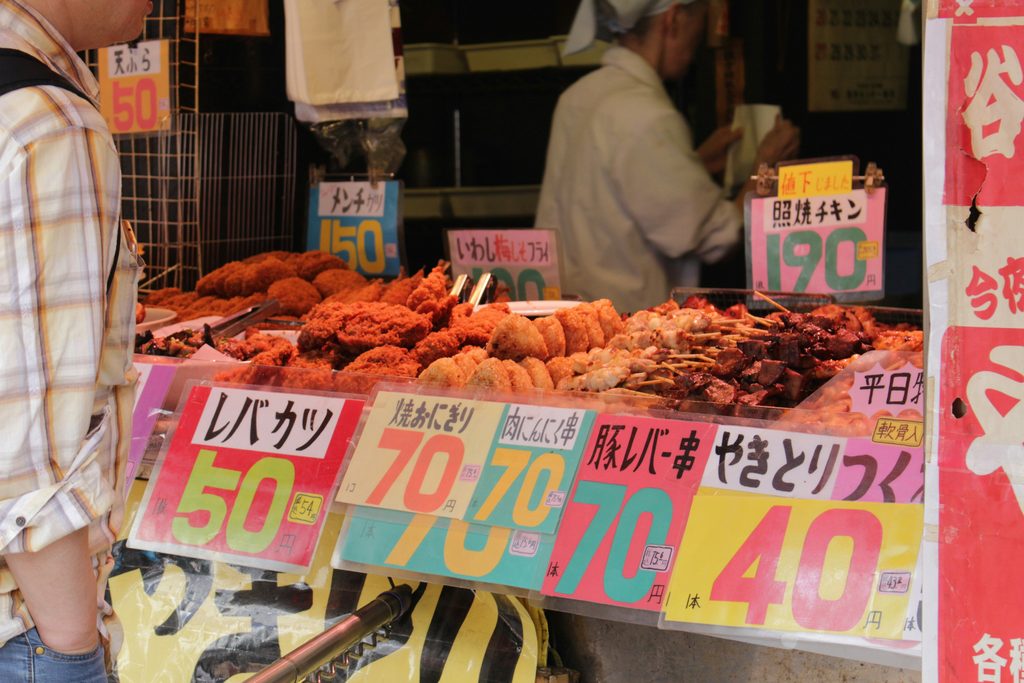 Japanese street food stall displaying fried snacks and grilled skewers with colorful handwritten price signs.